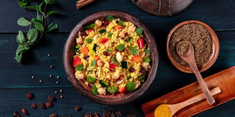 una ciotola piena di cous cous accanto a erbe aromatiche e utensili di legno