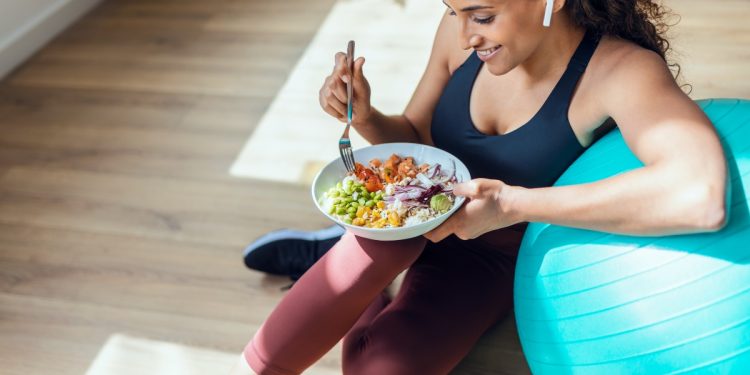 Donna in casa vestita da palestra, appoggiata su una fitball che mangia un insalata perché segue la dieta anticellulite