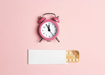su fondo rosa un orologio a sveglia rosa e sotto una confezione di pillole