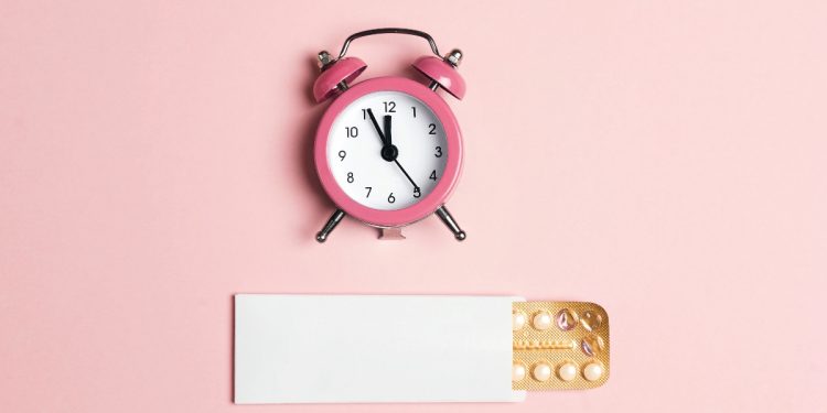 su fondo rosa un orologio a sveglia rosa e sotto una confezione di pillole