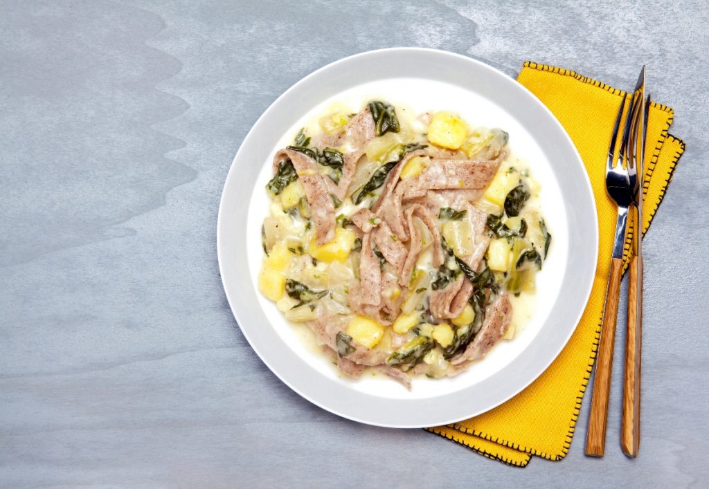 Pizzoccheri, la ricetta classica da preparare a casa 3 su piano di cucina grigio, piatto bianco con i pizzoccheri e accanto tovagliolo giallo forchetta e coltello