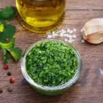 su tavolo di legno, ciotola di vetro con salsa verde alla Piemontese e attorno ingredienti come aglio, olio EVO, pepe e sale