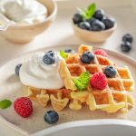 Waffle senza burro, la ricetta leggera per una colazione sana e golosa 1 su base chiara piatto piano con waffles e frutti di bosco, ciotola con yogurt e ciotola con mirtilli accanto