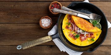 su tavolo di legno, padella con un omelette pronta con forchetta e coltello e accanto sale e pepe