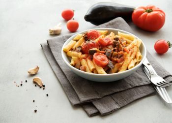 su tavolo bianco un tovagliolo grigio con sopra un piatto bianco con pasta con melanzane e pomodorini. A destra, forchetta e coltelli messi alla rovescia e dietro pomodori e melanzana