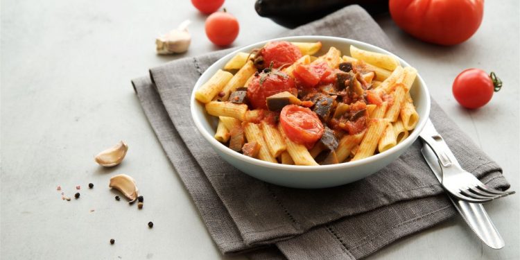 su tavolo bianco un tovagliolo grigio con sopra un piatto bianco con pasta con melanzane e pomodorini. A destra, forchetta e coltelli messi alla rovescia e dietro pomodori e melanzana