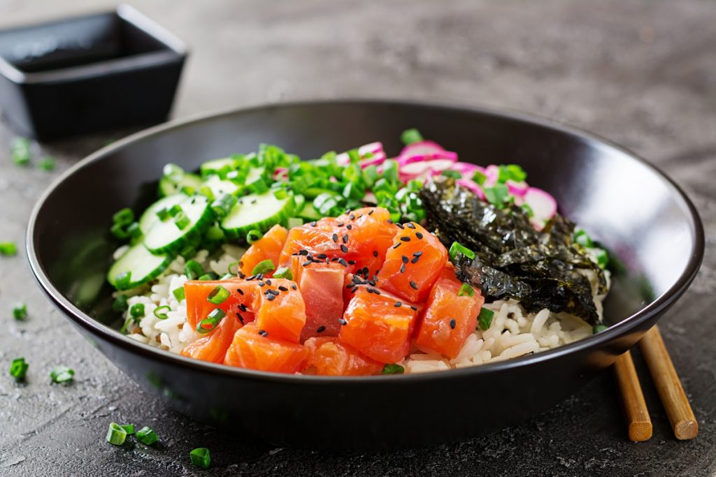 su tavolo di marmo: ciotola con poke bowl con alghe e salmone. Accanto bacchette cinese e dietro ciottola piccola con salsa di soia