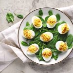 un piatto tondo su un canovaccio bianco con delle uova ripiene tagliate a metà sopra foglie si spinaci