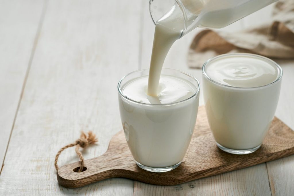 Yogurt fatto in casa, una ricetta semplice con solo 2 ingredienti 2 su tavolo di legno bianco, tagliere di legno con 2 bicchieri con yogurt fatto in casa e una mano che versa il latte sopra