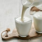 su tavolo di legno bianco, tagliere di legno con 2 bicchieri con yogurt fatto in casa e una mano che versa il latte sopra