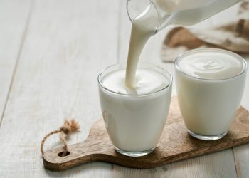 su tavolo di legno bianco, tagliere di legno con 2 bicchieri con yogurt fatto in casa e una mano che versa il latte sopra