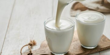 su tavolo di legno bianco, tagliere di legno con 2 bicchieri con yogurt fatto in casa e una mano che versa il latte sopra