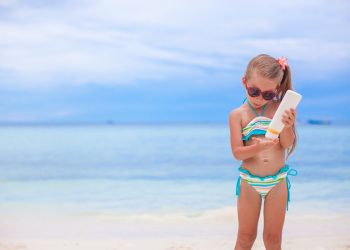 bambina sulla spiaggia con occhiali da sole e una crema solare in mano