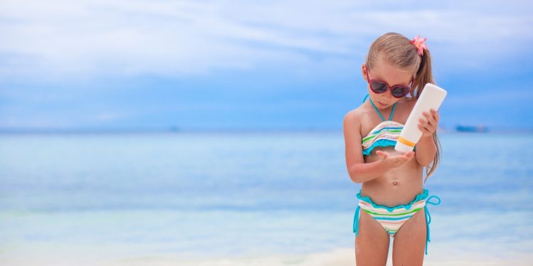 bambina sulla spiaggia con occhiali da sole e una crema solare in mano