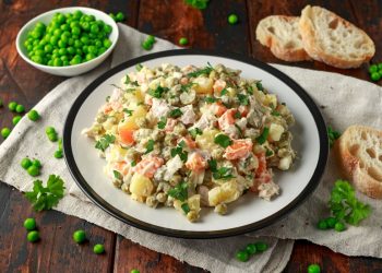 su tavolo di legno sotto piatto bianco con piatto bianco con insalata russa, accanto piselli e fette di pane