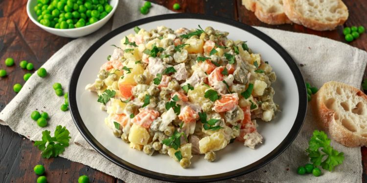 su tavolo di legno sotto piatto bianco con piatto bianco con insalata russa, accanto piselli e fette di pane