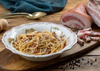 su tavolo di legno, tagliere di legno con un piatto di pasta alla Gricia e attorno del bacon tagliato, pepe nero e un cucchiaino