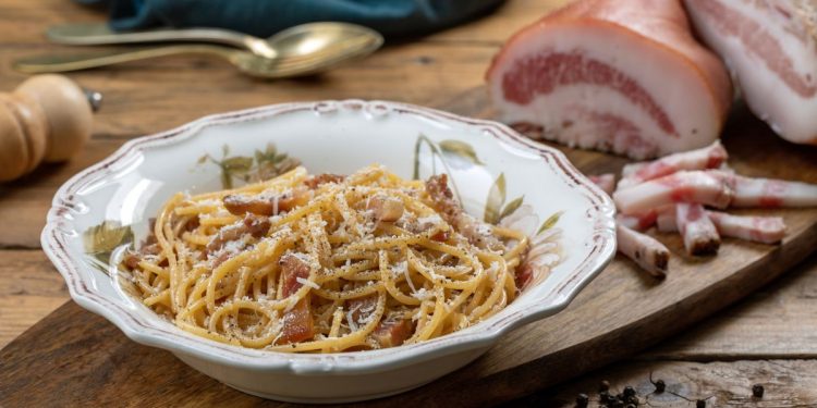 su tavolo di legno, tagliere di legno con un piatto di pasta alla Gricia e attorno del bacon tagliato, pepe nero e un cucchiaino