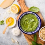 su piano di lavoro di marmo tagliere con una ciotola piena di pesto alla Genovese e attorno gli ingredienti, limone, basilico, aglio e basilico. Sul tagliere ciotola piena di pinoli