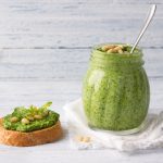 Pesto di rucola: la ricetta cremosa pronta in 10 minuti 1 In primo piano su uno sfondo chiaro, in un barattolo di vetro c'è un pesto di rucola con dei pinoli e un cucchiaino. Accanto al barattolo c'è una bruschetta con la crema di pesto.