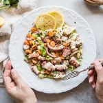 su tavolo bianco, canovaccio beige con piatto bianco con 2 mani in campo . La mano destra tiene la forchetta e sta per prendere un pò di insalata di polpo con carote e sedano. Attorno limone tagliato in 2.