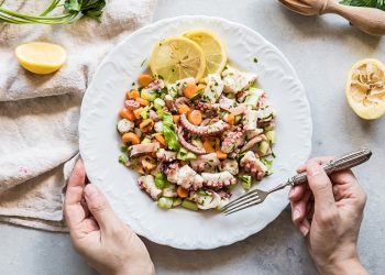su tavolo bianco, canovaccio beige con piatto bianco con 2 mani in campo . La mano destra tiene la forchetta e sta per prendere un pò di insalata di polpo con carote e sedano. Attorno limone tagliato in 2.
