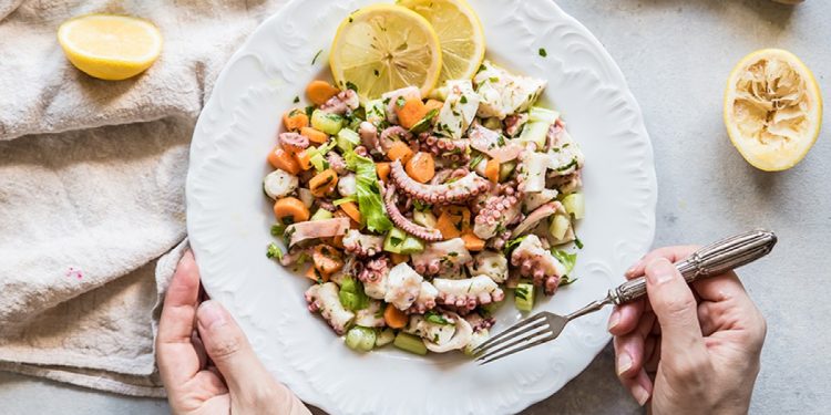 su tavolo bianco, canovaccio beige con piatto bianco con 2 mani in campo . La mano destra tiene la forchetta e sta per prendere un pò di insalata di polpo con carote e sedano. Attorno limone tagliato in 2.