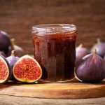 in primo piano su un tavolo di legno, un barattolo di vetro con la marmellata di fichi e dei fichi tagliati a metà intorno.