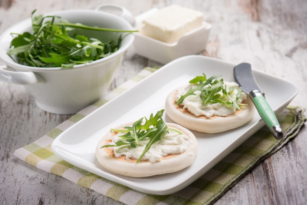 su tavolo di legno, tovagliolo a quadretti bianco e verdi con piatto bianco rettangolare e 2 tigelle di Modena con ricotta e stracchino . Accanto contenitore con rucola e in un'altro stracchino