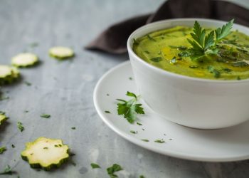 su base grigia piattino e tazza da brodo bianca piena di vellutata di zucchine. Rondelle di zucchine sparse