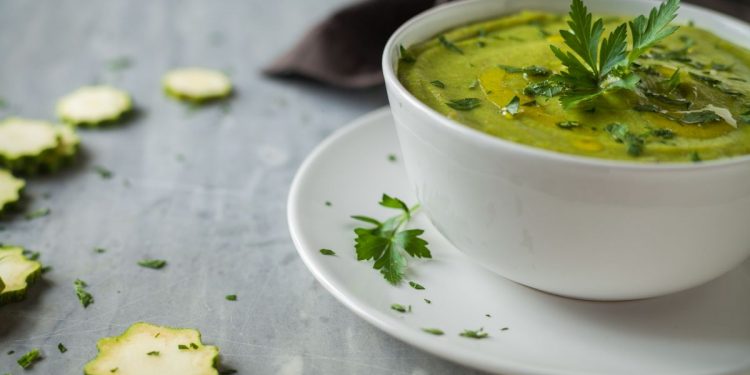 su base grigia piattino e tazza da brodo bianca piena di vellutata di zucchine. Rondelle di zucchine sparse