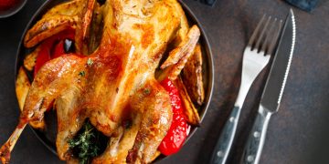 Primo piano di una piatto nero su tavolo da cucina con una faraona al forno, accanto forchetta e coltello