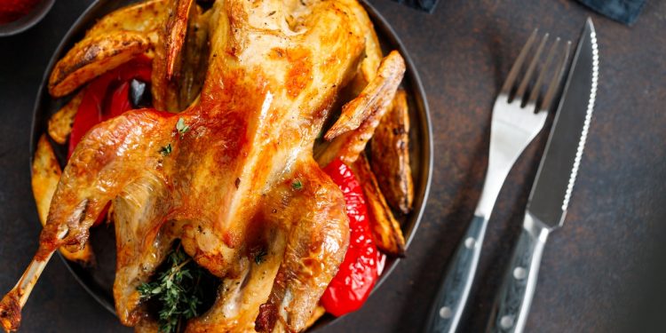 Primo piano di una piatto nero su tavolo da cucina con una faraona al forno, accanto forchetta e coltello