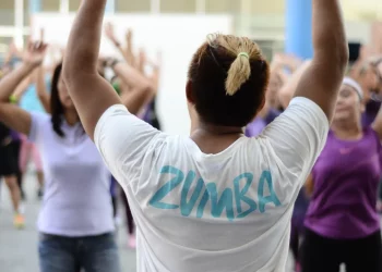 In una palestra, una lezione con un insegnante di schiena che indossa una maglietta bianca con la scritta zumba