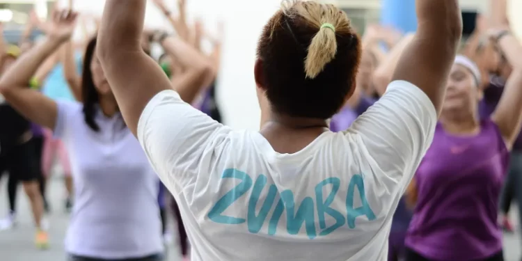 In una palestra, una lezione con un insegnante di schiena che indossa una maglietta bianca con la scritta zumba