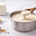 In primo piano su un tavolo chiaro c'è un pentola con la besciamella, sullo sfondo gli ingredienti della ricetta come noce moscata, burro e latte.