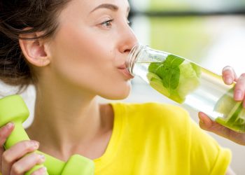 una donna con maglietta gialla e due piccoli pesi in una mano, beve una bevanda al lime con l'altra mano