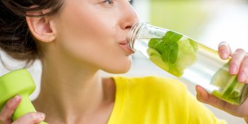 una donna con maglietta gialla e due piccoli pesi in una mano, beve una bevanda al lime con l'altra mano
