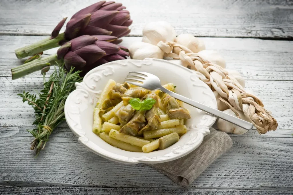 pasta con i carciofi: su tavolo di legno piatto bianco con pasta con i carciofi e forchetta appoggiata, sulla sinistra 2 carciofi , un rame di erbe aromatiche e a destra aglio