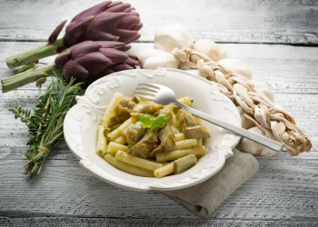 pasta con i carciofi: su tavolo di legno piatto bianco con pasta con i carciofi e forchetta appoggiata, sulla sinistra 2 carciofi , un rame di erbe aromatiche e a destra aglio