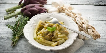 pasta con i carciofi: su tavolo di legno piatto bianco con pasta con i carciofi e forchetta appoggiata, sulla sinistra 2 carciofi , un rame di erbe aromatiche e a destra aglio