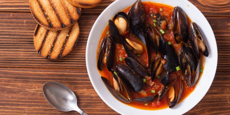 piatto bianco su un tavolo di legno con una zuppa di cozze, un cucchiaio e 3 fette di pane tostato