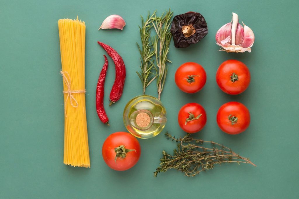 Pasta all'arrabbiata: la ricetta classica della tradizione 2 su sfondo verde gli ingredienti della pasta all'arrabbiata : spaghetti, peperoncino, aglio, pomodori, olio EVO, erbe aromatiche