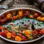 Orata all’acqua pazza: una ricetta di pesce leggera e saporita 1 In primo piano su una tovaglietta in materiale grezzo c'è una pirofila in acciaio con l'orata all'acqua pazza e pomodorini. Sullo sfondo delle posate e una macina pepe.