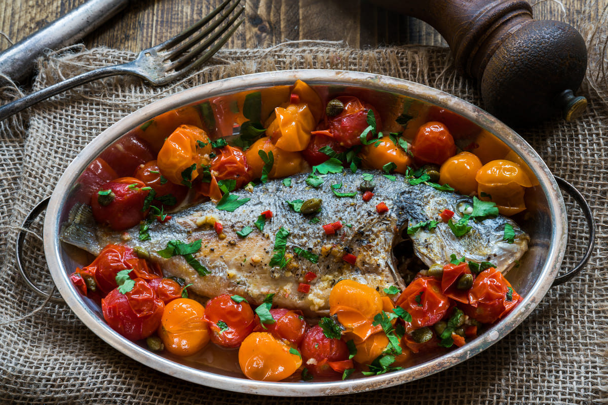 In primo piano su una tovaglietta in materiale grezzo c'è una pirofila in acciaio con l'orata all'acqua pazza e pomodorini. Sullo sfondo delle posate e una macina pepe.
