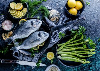 orata: su base grigia una padella con manico con dentro due orate, intorno una scodella di asparagi e fate di limone