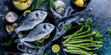 orata: su base grigia una padella con manico con dentro due orate, intorno una scodella di asparagi e fate di limone
