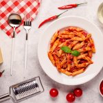 Pasta all'arrabbiata: la ricetta classica della tradizione 1 Su tavolo di marmo bianco, piatto con la pasta all'arrabbiata ( penne all'arrabbiata) e attorno gli ingredienti come pomodori e sugo di pomodori, peperoncino, olio EVO, parmigiano e la grattugia, una forchetta e un cucchiaio, postai su un canovaccio rosso a riga bianco