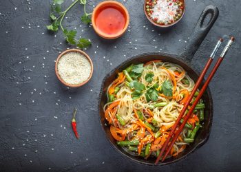 su base grigio scuro una padella con manico piena di spaghetti di riso con verdure, tre ciotole con salse varie, un paio di bacchette