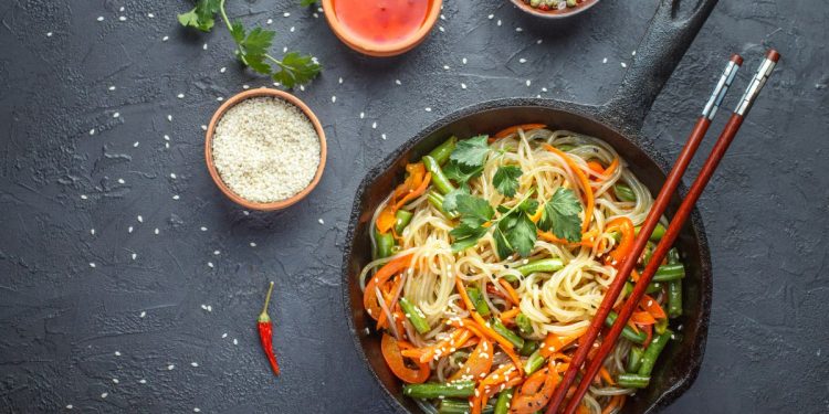 su base grigio scuro una padella con manico piena di spaghetti di riso con verdure, tre ciotole con salse varie, un paio di bacchette